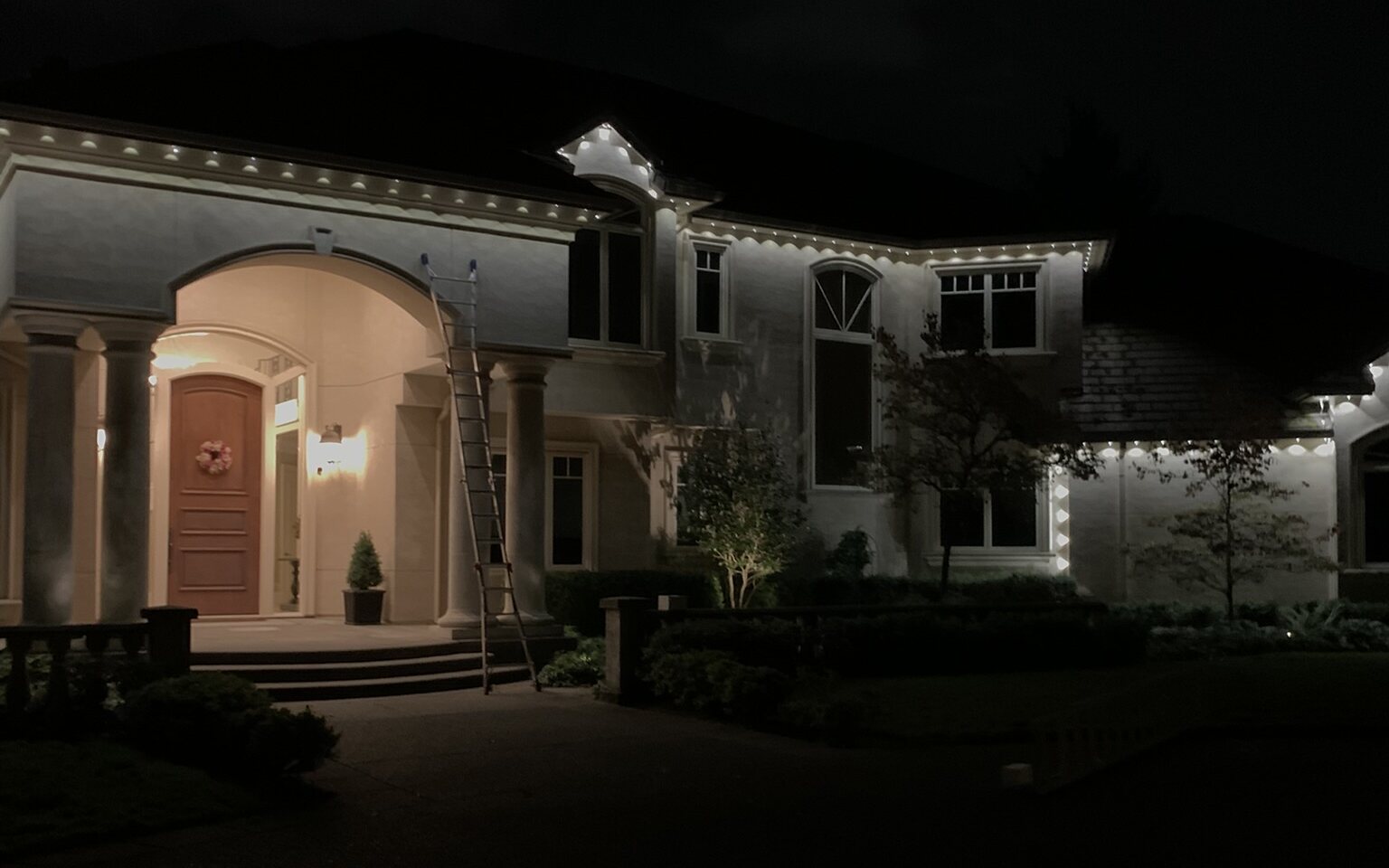 Large two-story stucco home at night with white holiday lights outlining the roofline and eaves, and a lit arched front entry